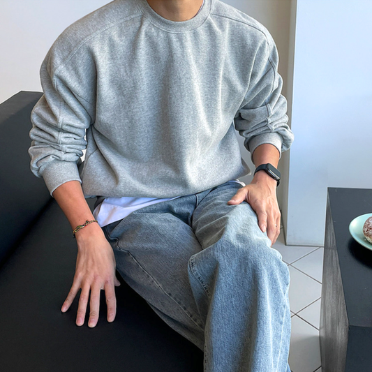 ライトグレーのショルダー切り替えクルーネックスウェット。ソファに座って片手を膝に置き、身頃と袖のゆとりを正面から見せた姿