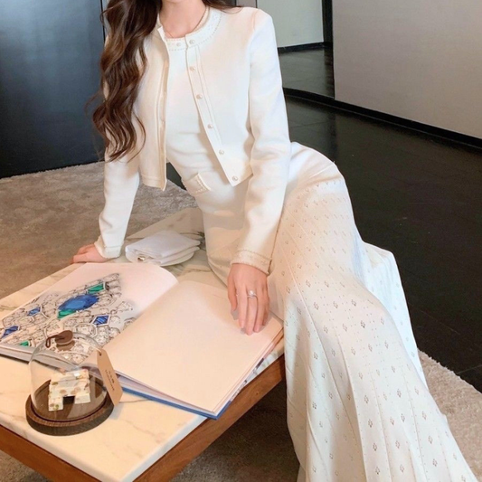 アイボリーのパール風装飾ニットワンピースとカーディガンのセット。カーディガンを羽織ってテーブル脇に腰掛けた姿。フロントのボタンと袖口の飾り