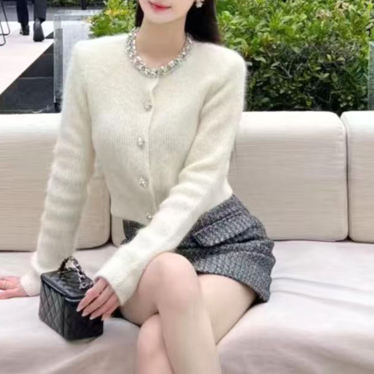 ホワイトのネックビジュー付きショート丈ニットカーディガン。女性がベンチに座った正面の上半身。スカートに重なる短めの裾と手元