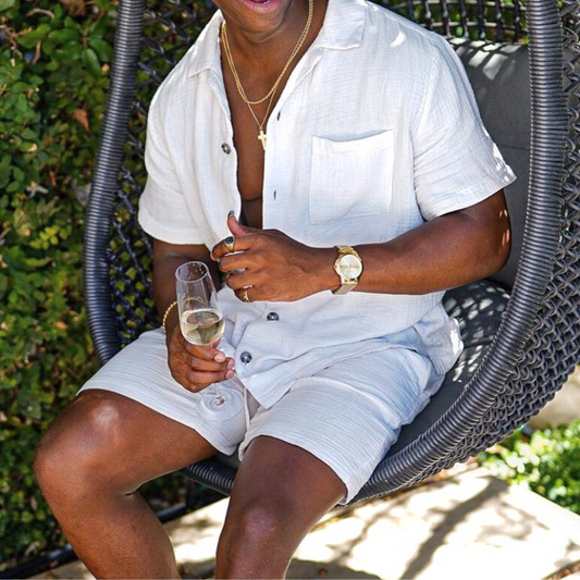 リネン風のホワイト開襟シャツとハーフパンツのセットアップを着た男性が屋外チェアでリラックスしている様子