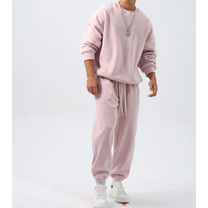ピンクのクルーネックトップスと裾絞りワイドパンツのフリースセットアップを着た男性。袖をたくし上げ、両腕を下げて白いスニーカーを合わせた正面の全身姿
