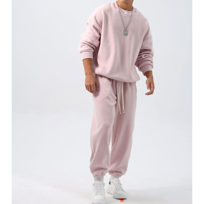 ピンクのクルーネックトップスと裾絞りワイドパンツのフリースセットアップを着た男性。袖をたくし上げ、両腕を下げて白いスニーカーを合わせた正面の全身姿