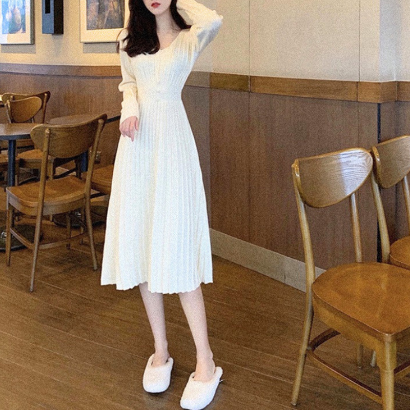 アイボリーのVネックプリーツリブニットワンピース。椅子の間で片手を頭に添えて立つ正面の全身。腰の切り替えから裾へ広がるAライン