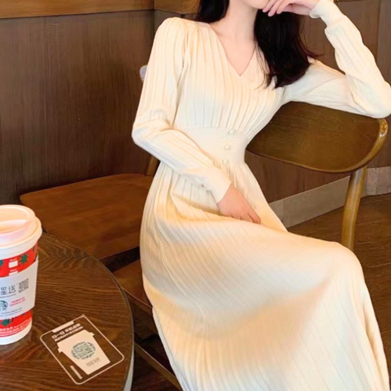 アイボリーのVネックプリーツリブニットワンピース。椅子に座り片手を頭に添えた正面の姿。胸元のVネックとウエストの小さなボタン