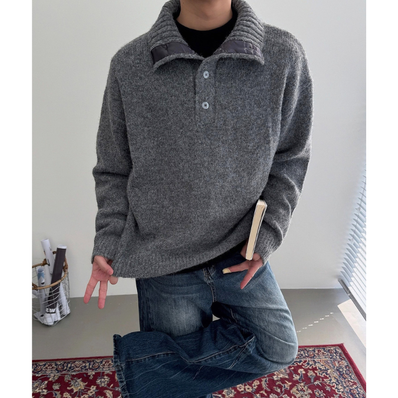 グレーのボタン付きモックネックニットプルオーバーを着た男性の上半身。襟を開き、片脚を曲げて本を持った姿