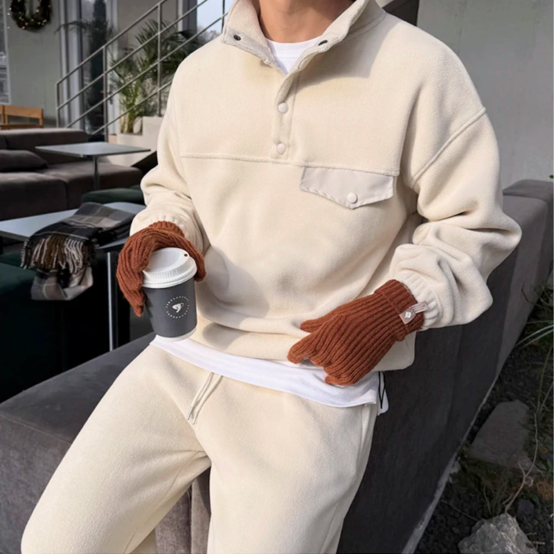 アイボリーのスタンドカラープルオーバーとワイドパンツのセットアップを着た男性。ソファの縁に寄りかかり、両手にカップと茶色の手袋を持つ上半身