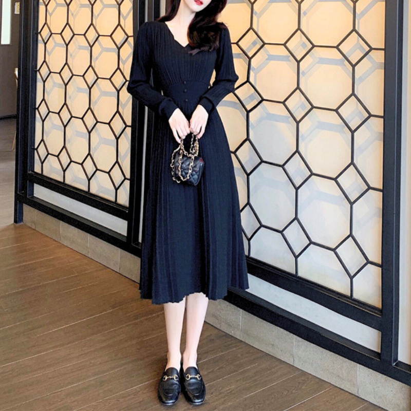 ブラックのVネックプリーツリブニットワンピース。幾何柄の仕切りの前でバッグを前に持って立つ正面の全身。足首が見える丈とAライン