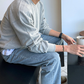 ライトグレーのショルダー切り替えクルーネックスウェット。ソファに座って白いカップを持ち、横から袖のふくらみと裾を写した姿