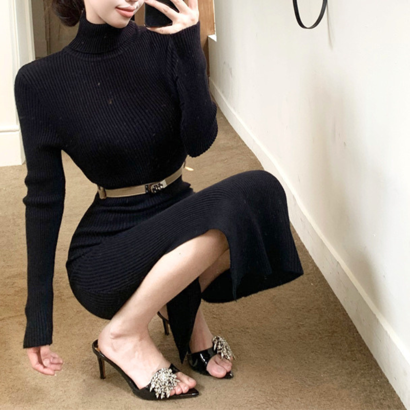 ブラックのベルト付きハイネックタイトニットワンピース。女性が腰を落として膝を曲げた着用姿。スリットから見える脚と黒い裾