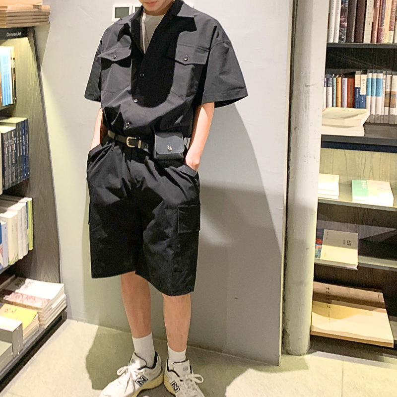 ブラックの半袖カーゴジャンプスーツを本棚の前で着た男性の立ち姿