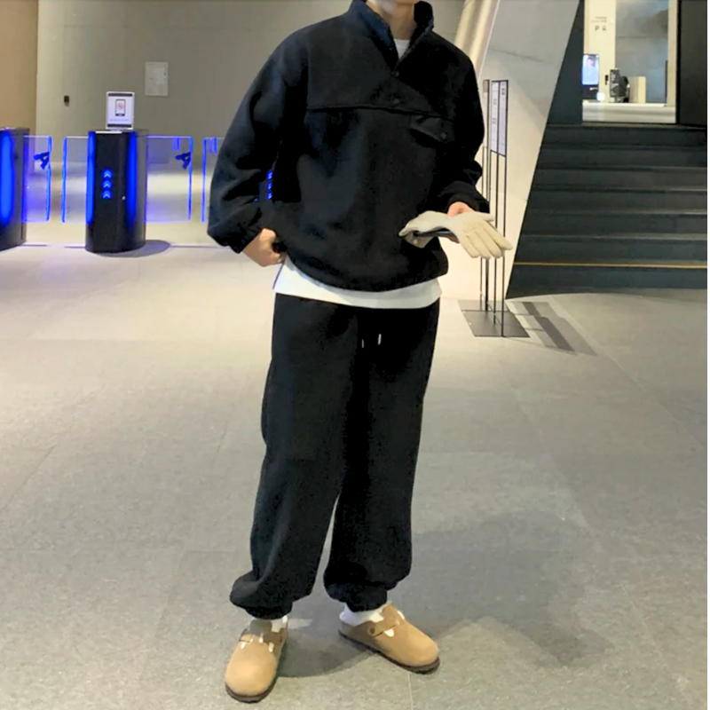 ブラックのスタンドカラープルオーバーとワイドパンツのセットアップを着た男性。正面に立ち、片方の白い手袋をもう片方の手で整えた全身姿