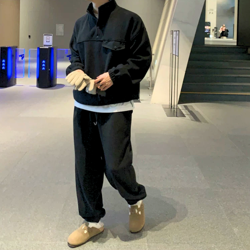 ブラックのスタンドカラープルオーバーとワイドパンツのセットアップを着た男性。白い手袋を両手で整えながら、脚を離して立った全身姿