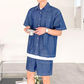 デニムライクな半袖シャツ×ショーツのセットアップを着た男性が室内でカメラを持って立つ姿