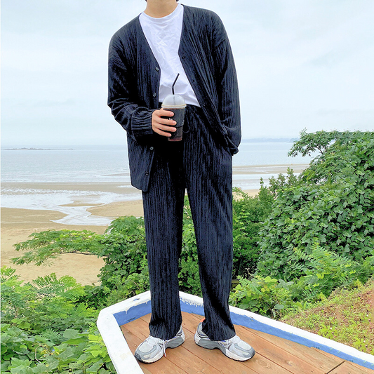 プリーツ風カーディガンセットアップを着た男性が海辺でコーヒーを手に立っている様子